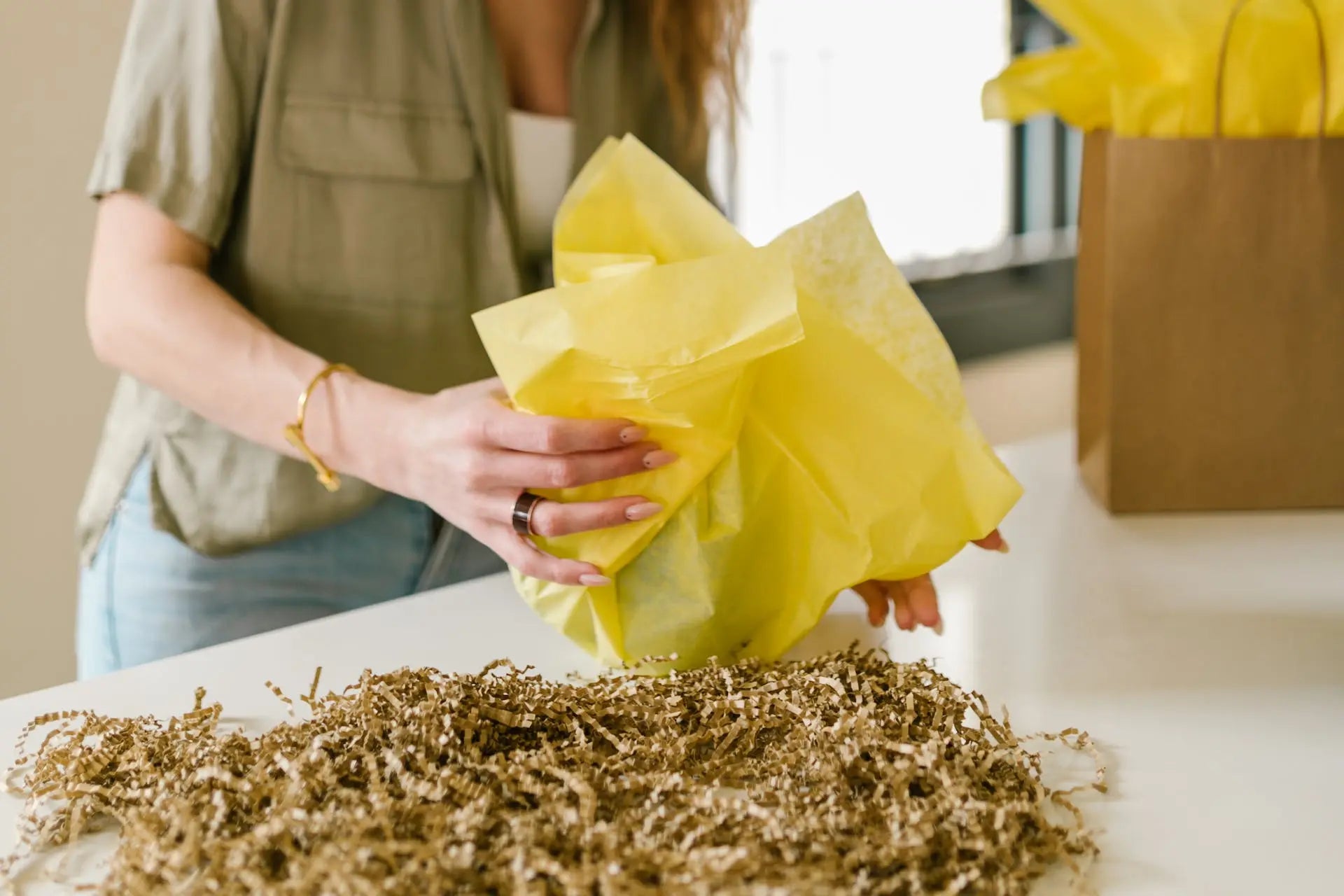 Person wrapping a gift with yellow acid-free tissue paper on a table with decorative filler.