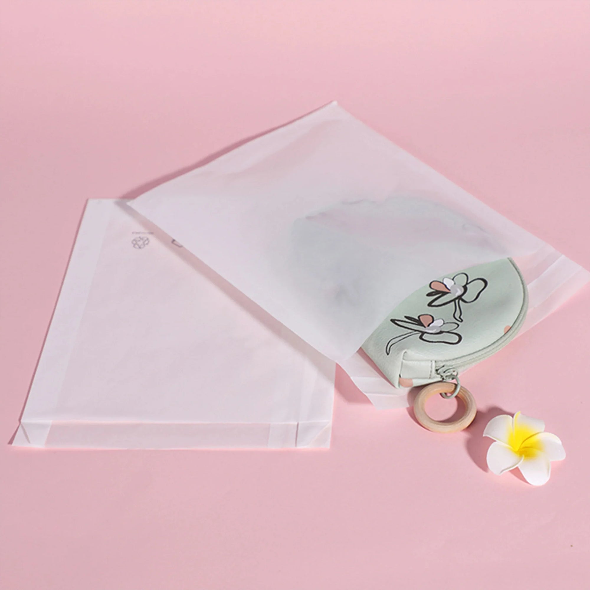 Glassine paper packaging with a green floral pouch and a plumeria flower on a pink background.