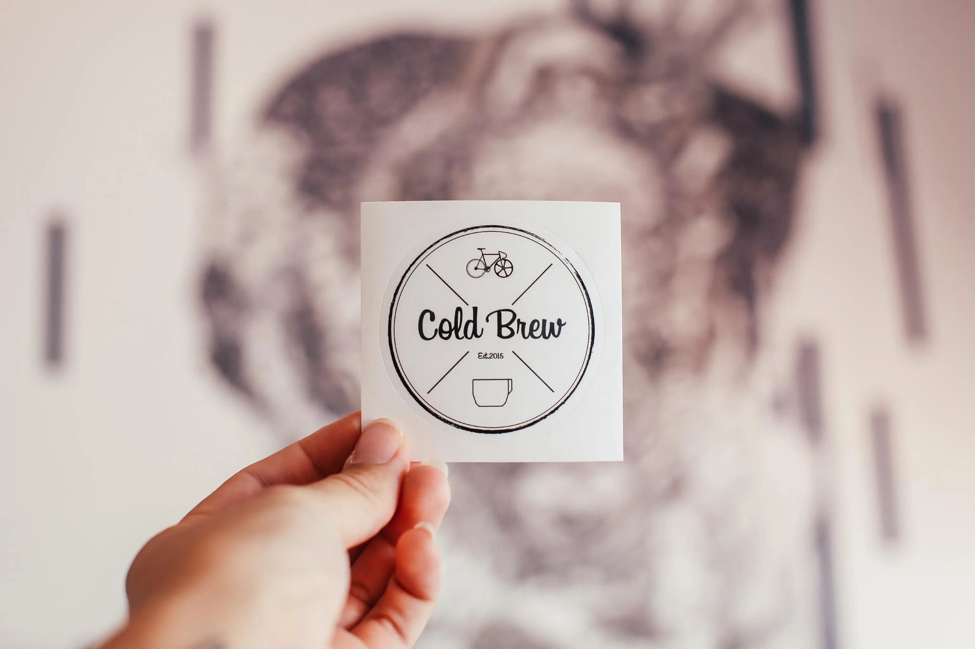 Hand holding a Cold Brew sticker featuring a bicycle and cup design against a blurred background.