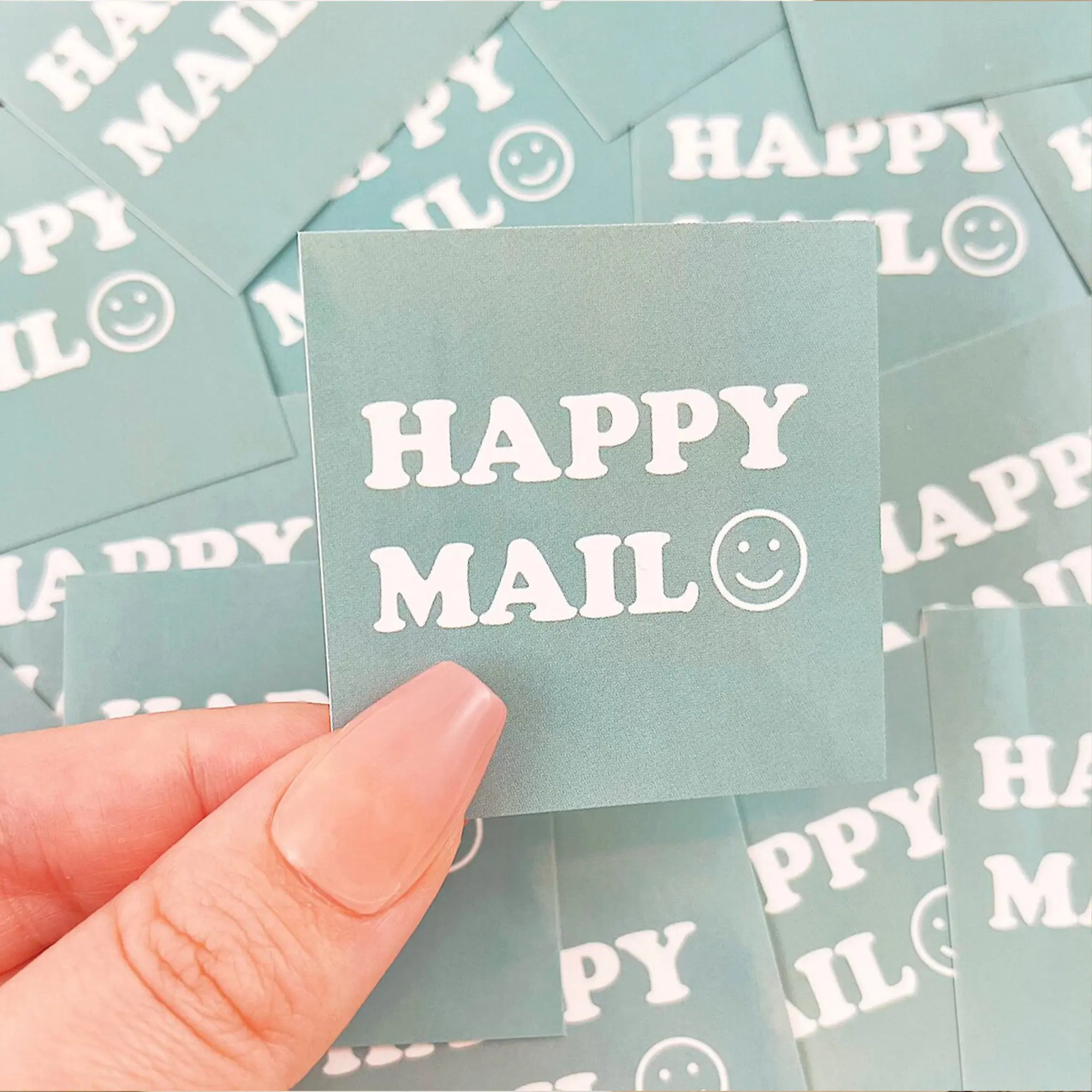 Hand holding a custom square sticker that reads "HAPPY MAIL" with a smiley face on a teal background.