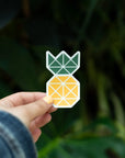 Custom die-cut pineapple sticker held by a person, featuring geometric design in green and yellow against a nature backdrop.