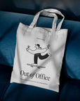 Custom tote bag with playful "Out of Office" design on a blue couch background.