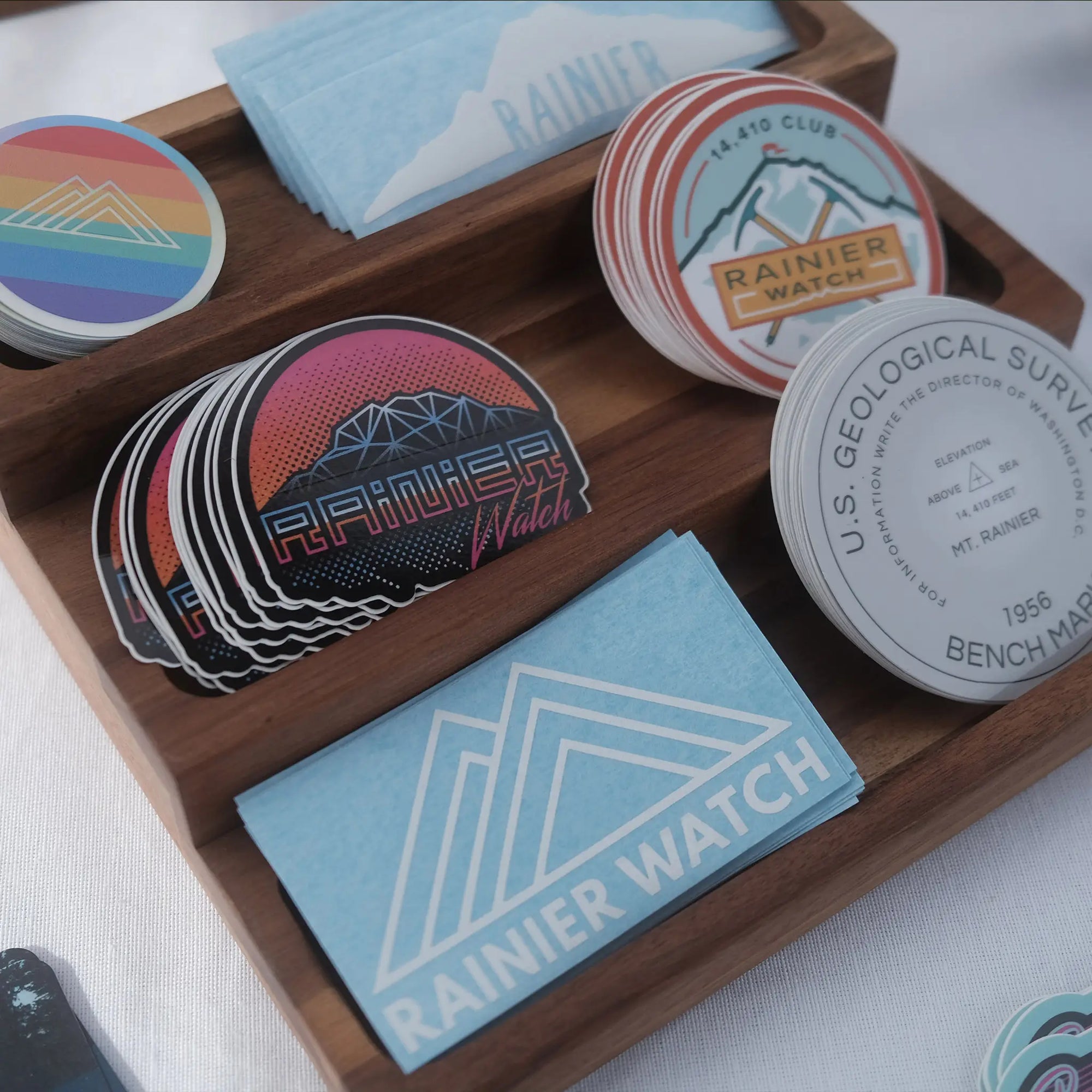 Custom die-cut stickers featuring various designs placed in a wooden display tray, promoting Rainier Watch branding.