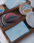 Custom die-cut stickers featuring various designs placed in a wooden display tray, promoting Rainier Watch branding.