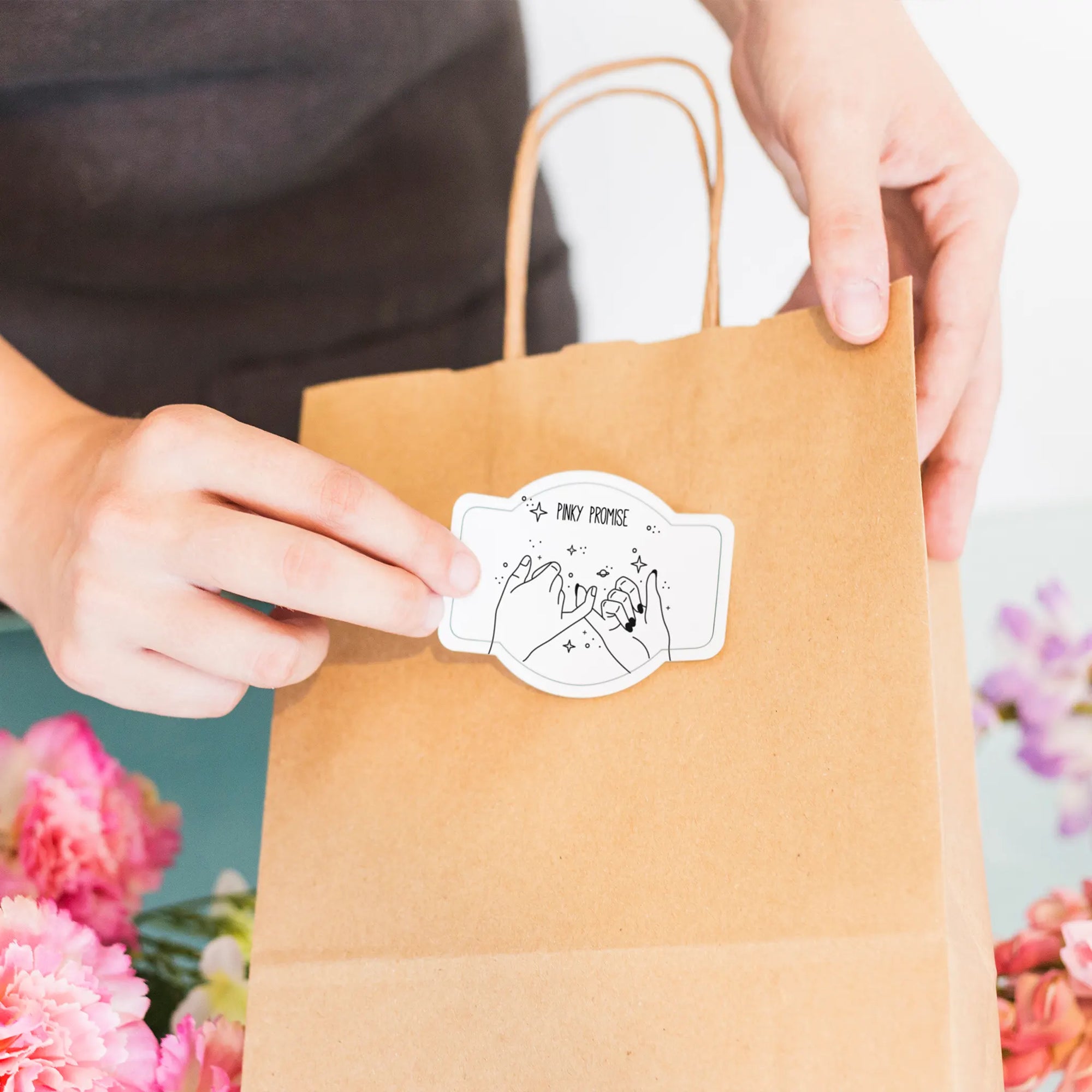 Person placing a custom die-cut sticker on a kraft paper bag, surrounded by flowers, showcasing personalization and creativity.