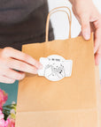 Person placing a custom die-cut sticker on a kraft paper bag, surrounded by flowers, showcasing personalization and creativity.