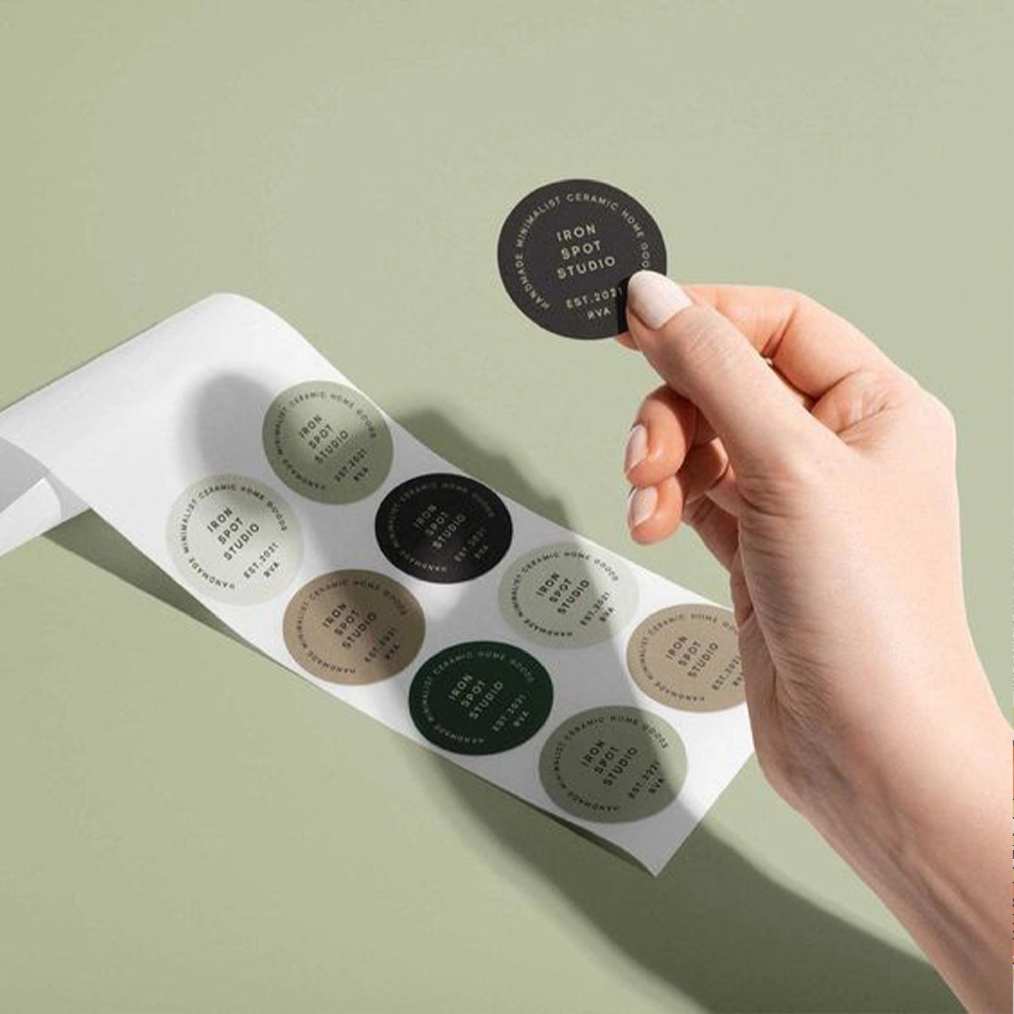 Hand holding a custom round sticker from a sheet on a green background, showcasing branding and design options.