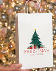 Custom white paper bag with Christmas tree design for gift shops, surrounded by festive holiday lights.