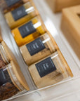 jars of spices and seasonings with wooden lids, arranged neatly in a clear container on a kitchen countertop
