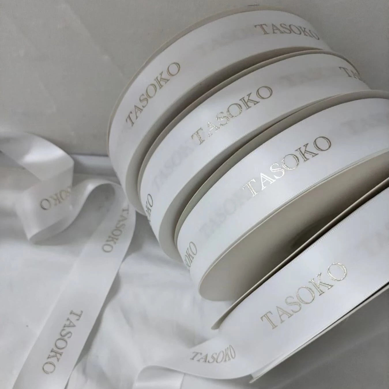 Custom satin ribbon rolls with embossed "TASOKO" logo, featuring a soft, luxurious finish. Perfect for gifts and decorations.
