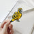 Custom die-cut yellow parrot sticker held above a notebook on a white background.