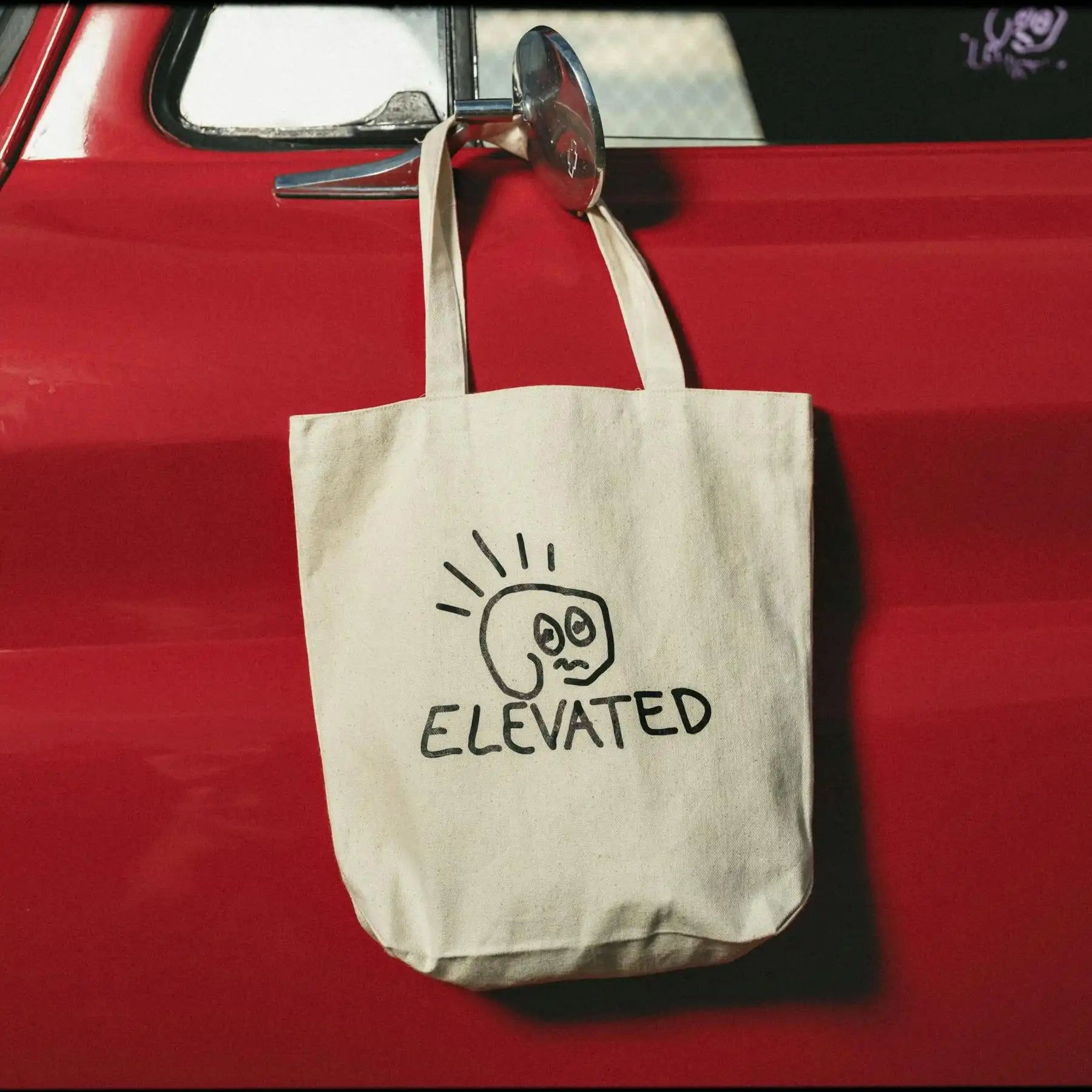 Custom tote bag with unique 'ELEVATED' design hanging on a red car door, showcasing style and sustainability.