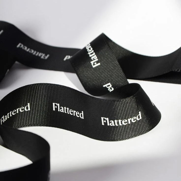 Custom black grosgrain ribbon with the word "Flattered" printed in white, showcasing its elegant texture and style.