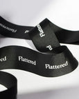 Custom black grosgrain ribbon with the word "Flattered" printed in white, showcasing its elegant texture and style.