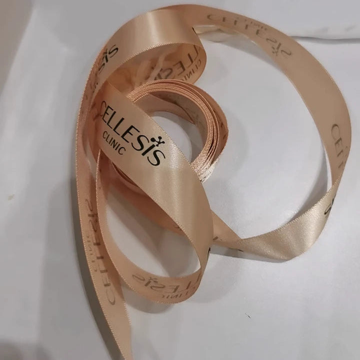 Custom satin ribbon in soft peach color with logo of Cellesis Clinic, elegantly coiled on a white surface.