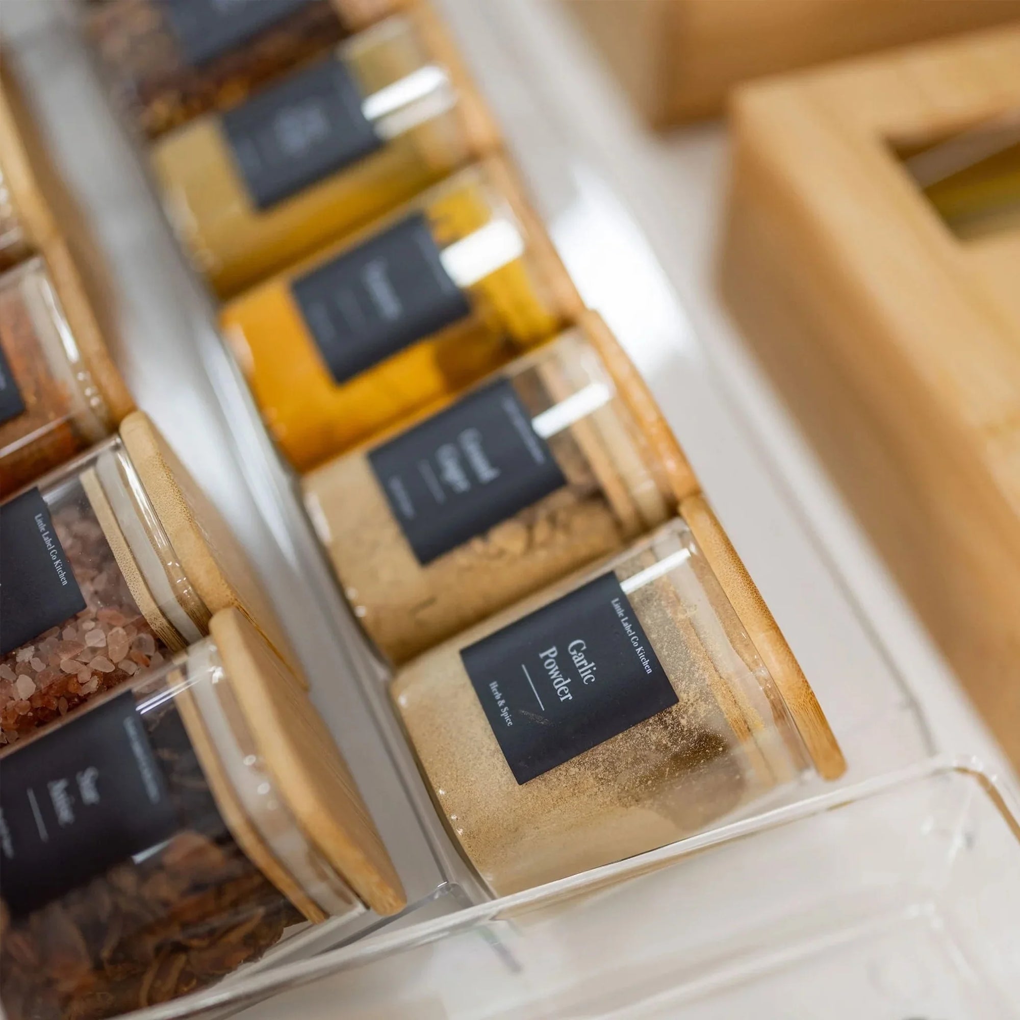 jars of spices and seasonings with wooden lids, arranged neatly in a clear container on a kitchen countertop
