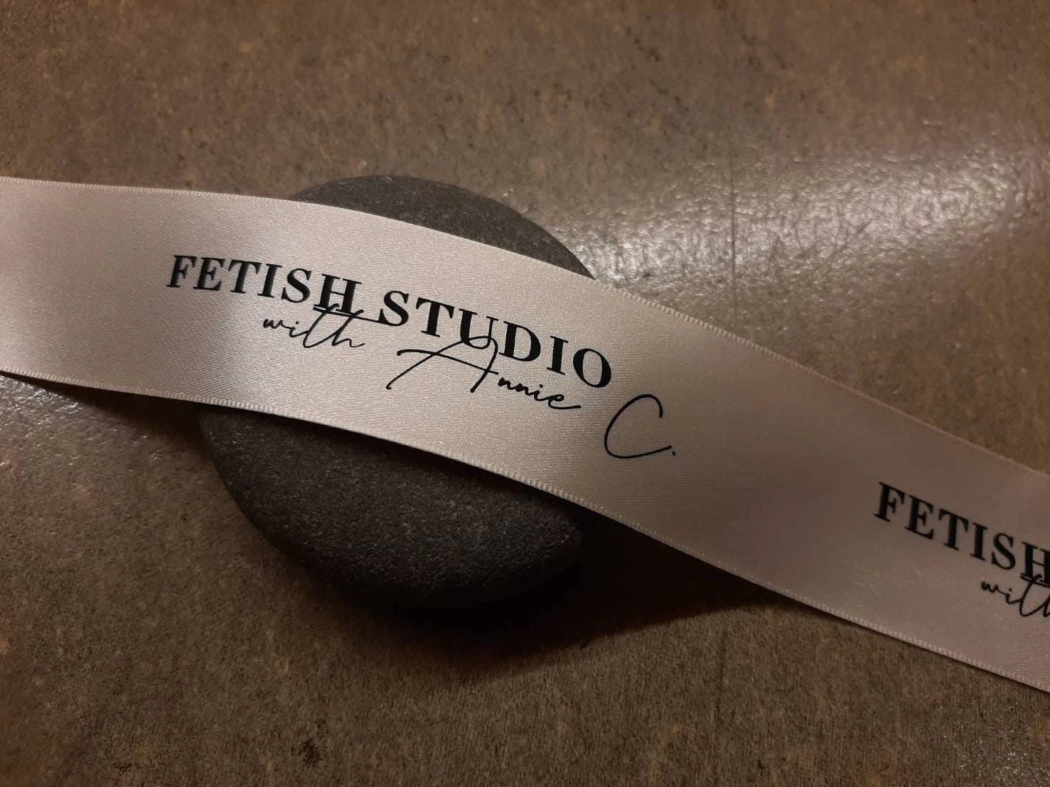 Custom satin ribbon featuring "Fetish Studio with Annie C" logo on a smooth stone background.